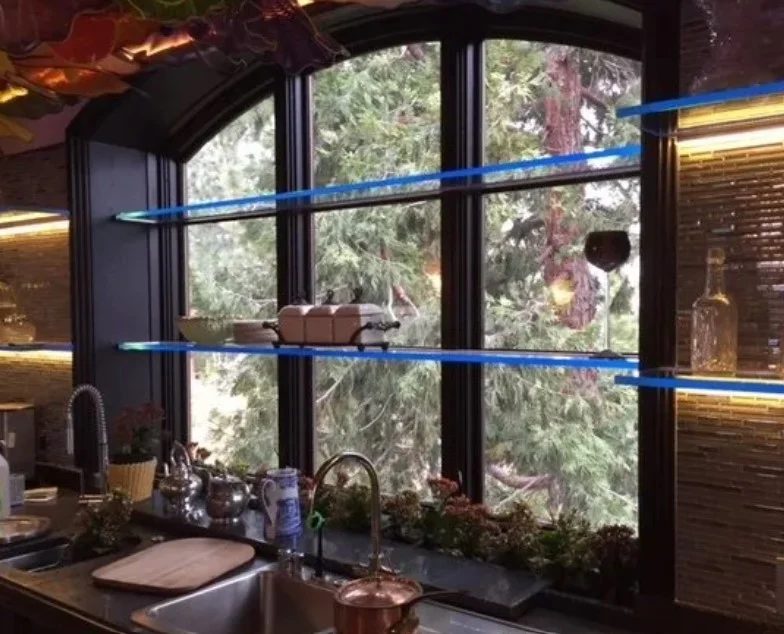 Edge-lit floating glass shelves spanning an arched kitchen window with Tahoe pines outside